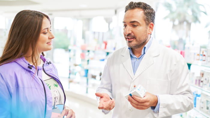 A pharmacist holding a small white box of medication consulting with a customer in a pharmacy. The medium-skinned pharmacist wears a white lab coat and the light-skinned customer wears a purple and blue jacket.